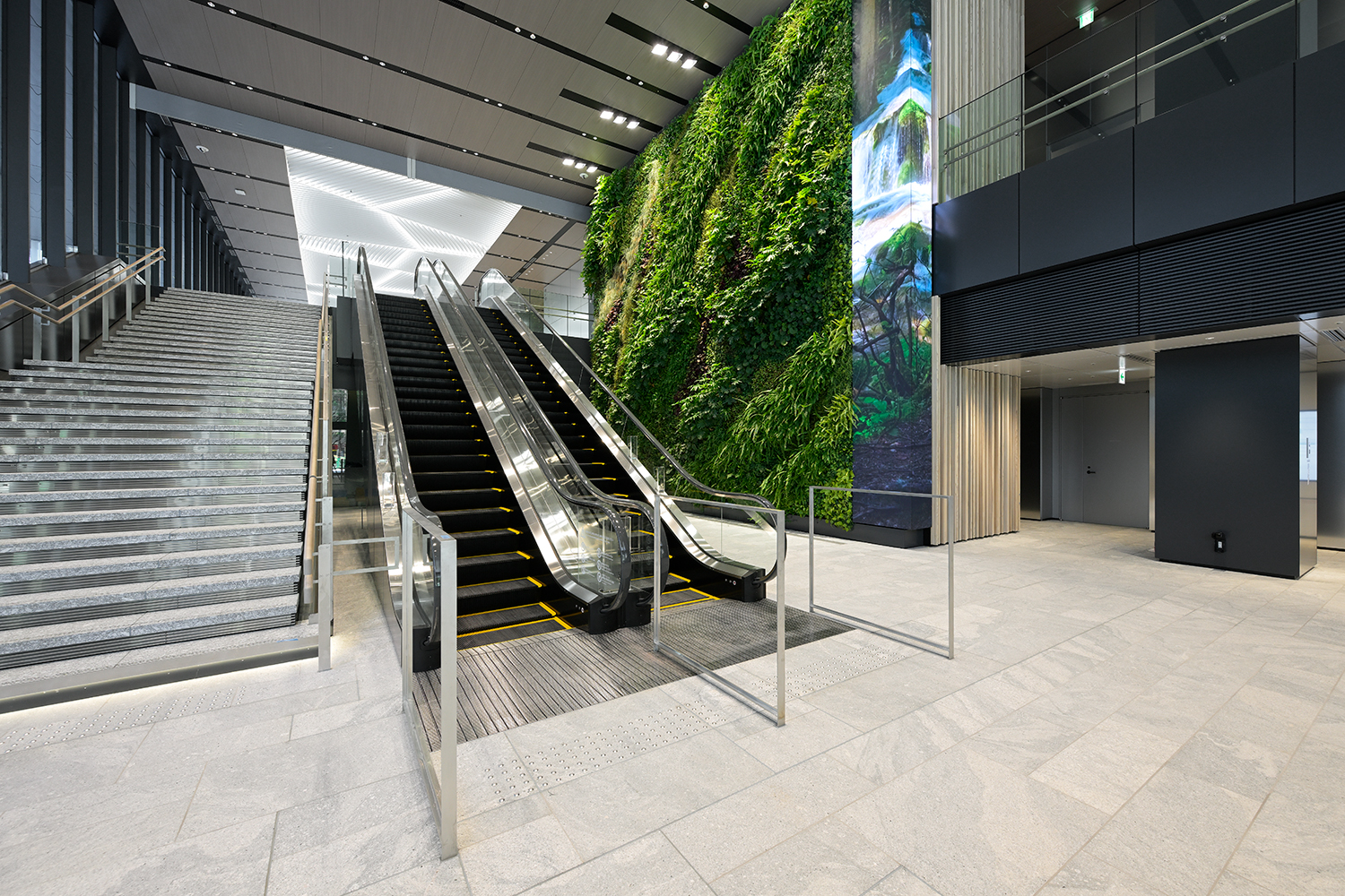 1階のエントランスホール。正面にエスカレーター、左手に階段を配置している。本物の植物をあしらい緑化された壁面（向かって右）と、地面から天面まで伸びるモニターが印象的（エスカレーター：No.A1，A2）